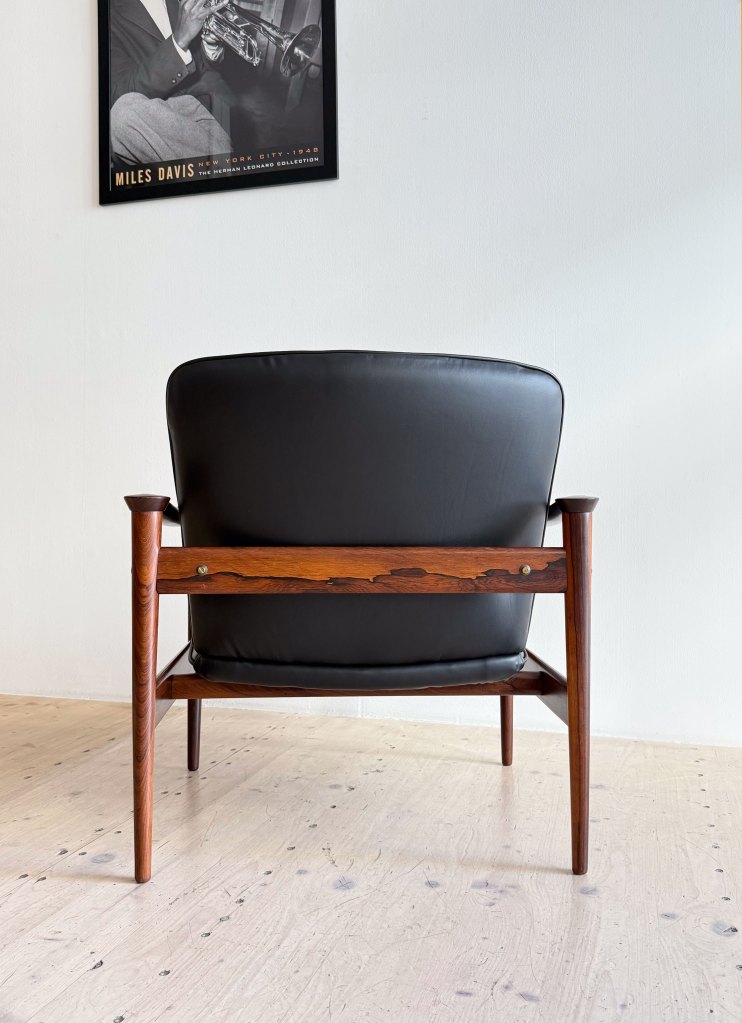 A mid-century modern chair with a black leather seat and wooden frame, viewed from the back. A framed poster of a musician is hung on the wall above.