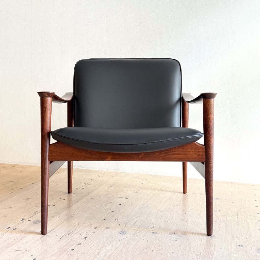 A modern armchair with a black leather seat and backrest, featuring wooden armrests and legs on a light wooden floor.