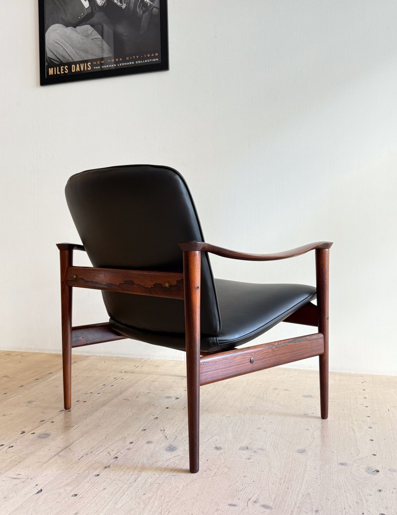 A mid-century modern wooden chair with a black leather seat, viewed from the back, set against a light-colored wall and wooden floor.