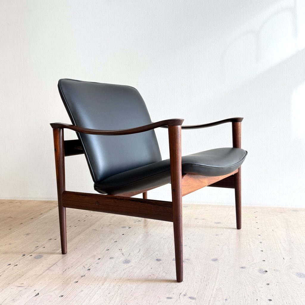 A modern chair with a sleek black leather seat and wooden frame, positioned on a light wooden floor against a plain wall.
