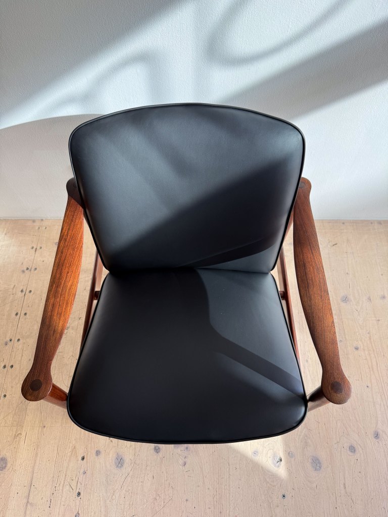 Top view of a modern black upholstered chair with wooden armrests on a light wood floor, illuminated by natural light.