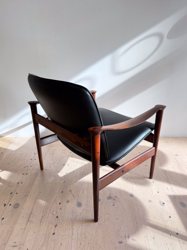 Elegant wooden chair with a black leather seat, viewed from the back, showcasing minimalist design and natural light casting shadows on the wall.
