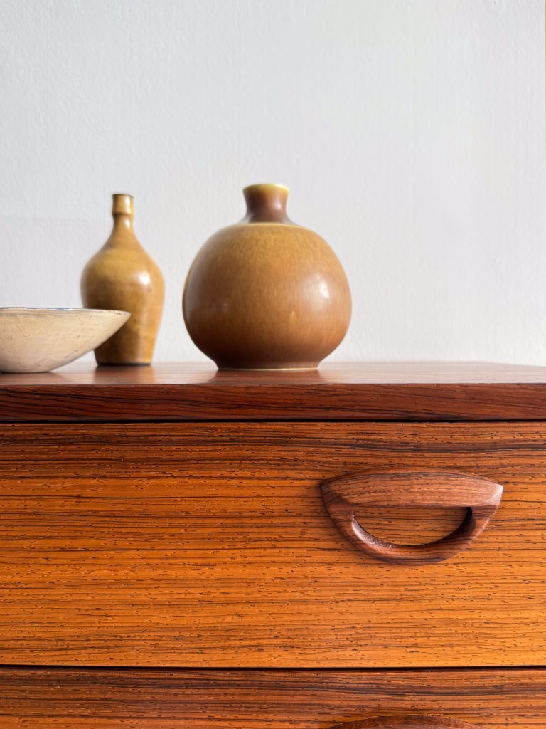 A wooden dresser with a textured surface, featuring a brown decorative bowl and two ceramic vases in shades of brown on top.