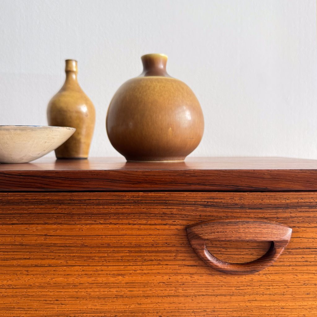 A close-up of a wooden dresser featuring a curved handle, with three decorative ceramic vases placed on top. The vases are in earthy tones, and there's a shallow bowl beside them, all set against a plain white wall.