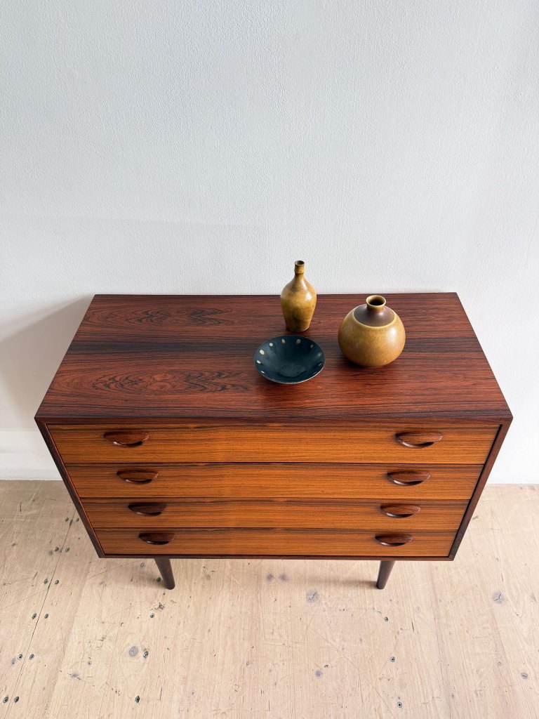 A wooden dresser with four drawers, featuring a rich brown finish and curved handles, topped with two decorative vases and a small plate.