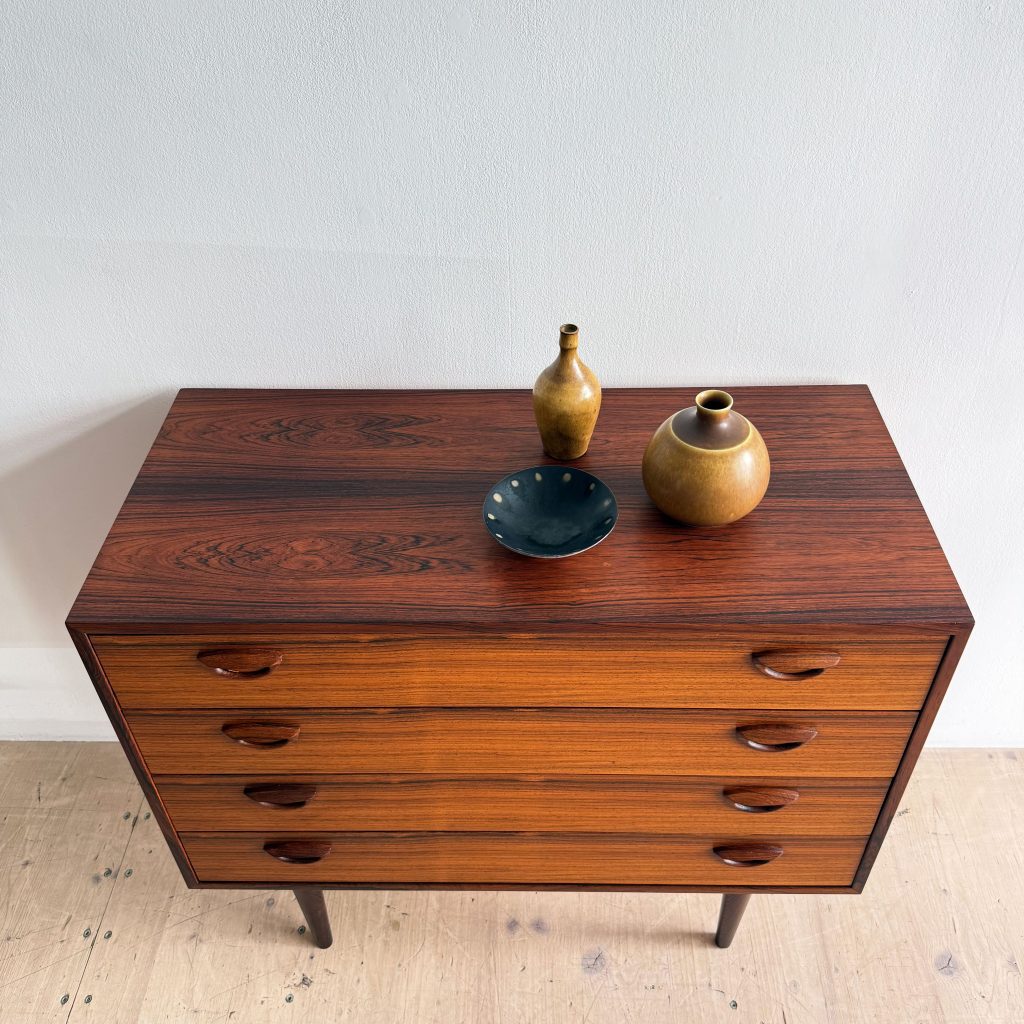 A top-down view of a wooden dresser with four drawers, featuring a smooth, dark finish. On top of the dresser, there are two ceramic vases, one brown and the other a darker shade, alongside a small green bowl with white polka dots.