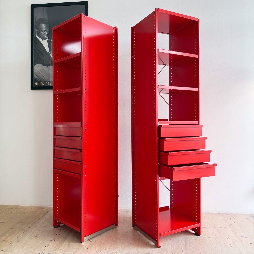 Two tall red storage units with shelves and drawers, set against a light-colored wall with a framed black and white portrait in the background.