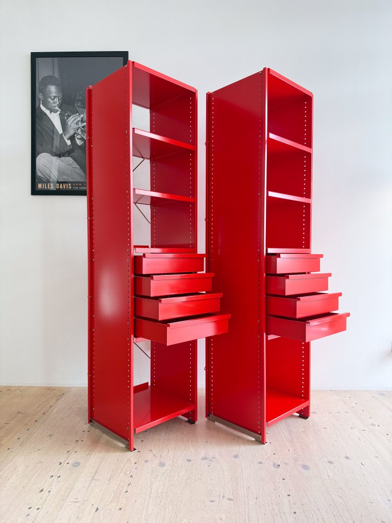 Two tall, modern red storage cabinets with open shelves and multiple drawers, set against a light-colored wall.