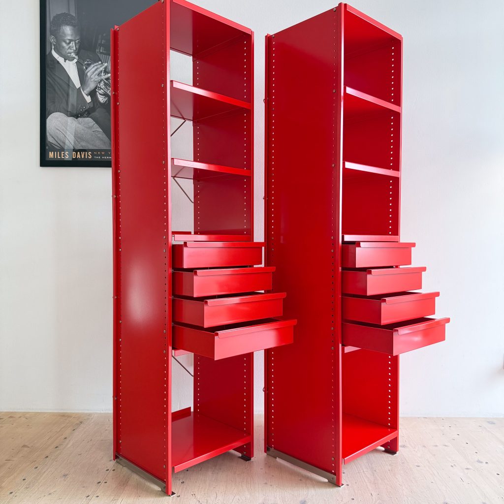 Two tall red storage units with open shelves and multiple drawers, set against a light wall.