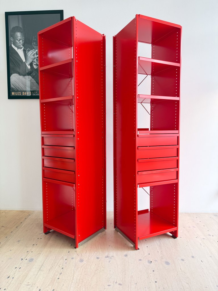 Two tall red shelving units with open shelves and drawers, placed against a white wall with a framed black and white photograph in the background.