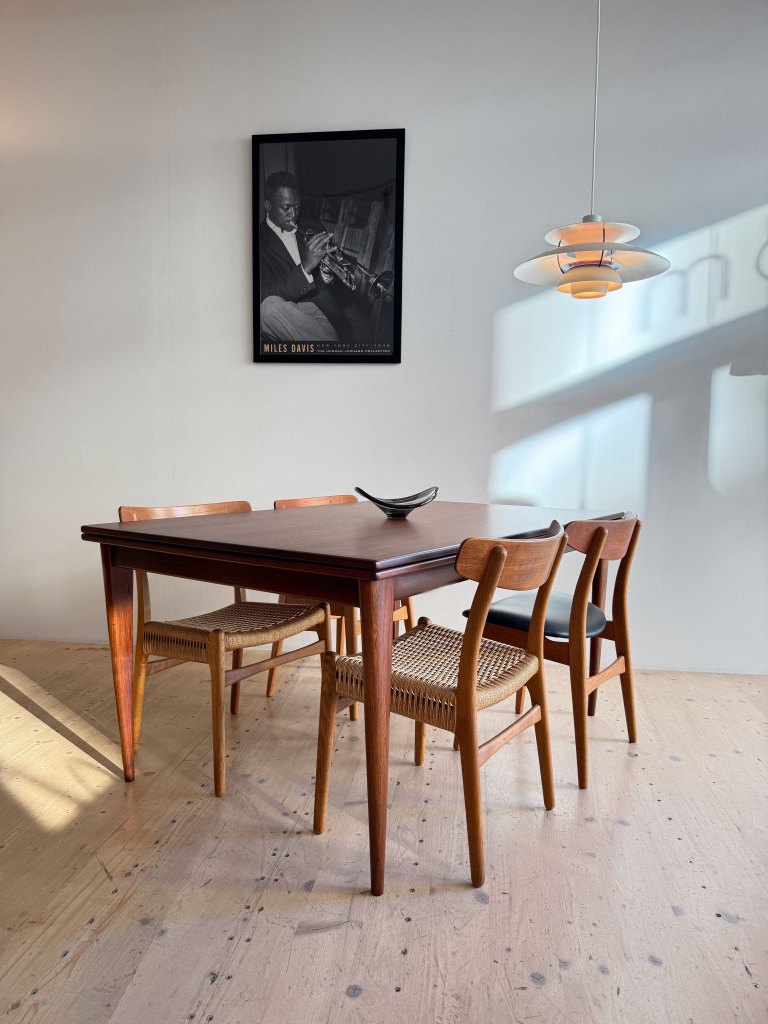 A stylish wooden dining table surrounded by four chairs with woven seats, featuring a decorative bowl on the table. A framed black and white portrait of Miles Davis hangs on the wall above the table, with a modern pendant light illuminating the scene.