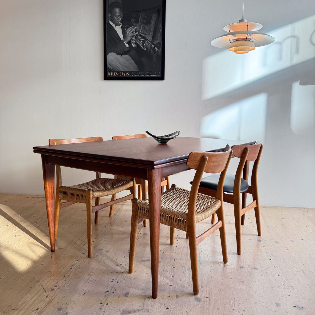 A modern wooden dining table surrounded by four chairs, featuring a minimalist design. A small decorative bowl is placed on the table, and a framed black and white poster of a musician hangs on the wall.
