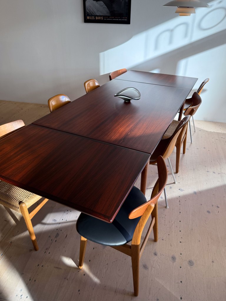 A modern wooden dining table with a sleek design and a decorative centerpiece, surrounded by various styles of chairs, illuminated by natural light.