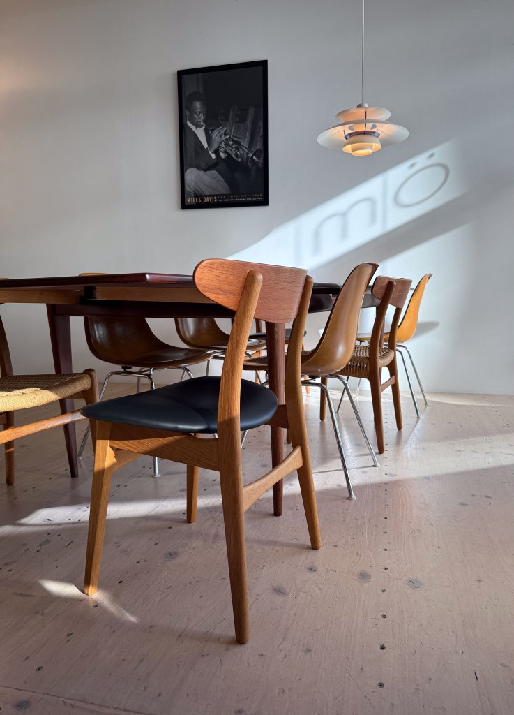 A modern dining room featuring a wooden table surrounded by several stylish chairs. A single pendant light hangs from the ceiling, and a black-and-white poster of a musician is visible on the wall. Soft shadows are cast on the floor.