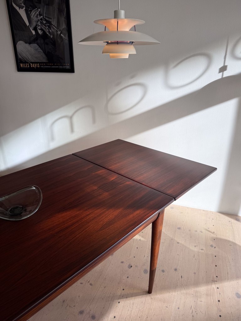 A wooden dining table with a glossy surface and tapered legs, featuring a glass decorative piece on top. A modern pendant light hangs above, casting shadows on the wall, and a framed poster of a musician is visible in the background.