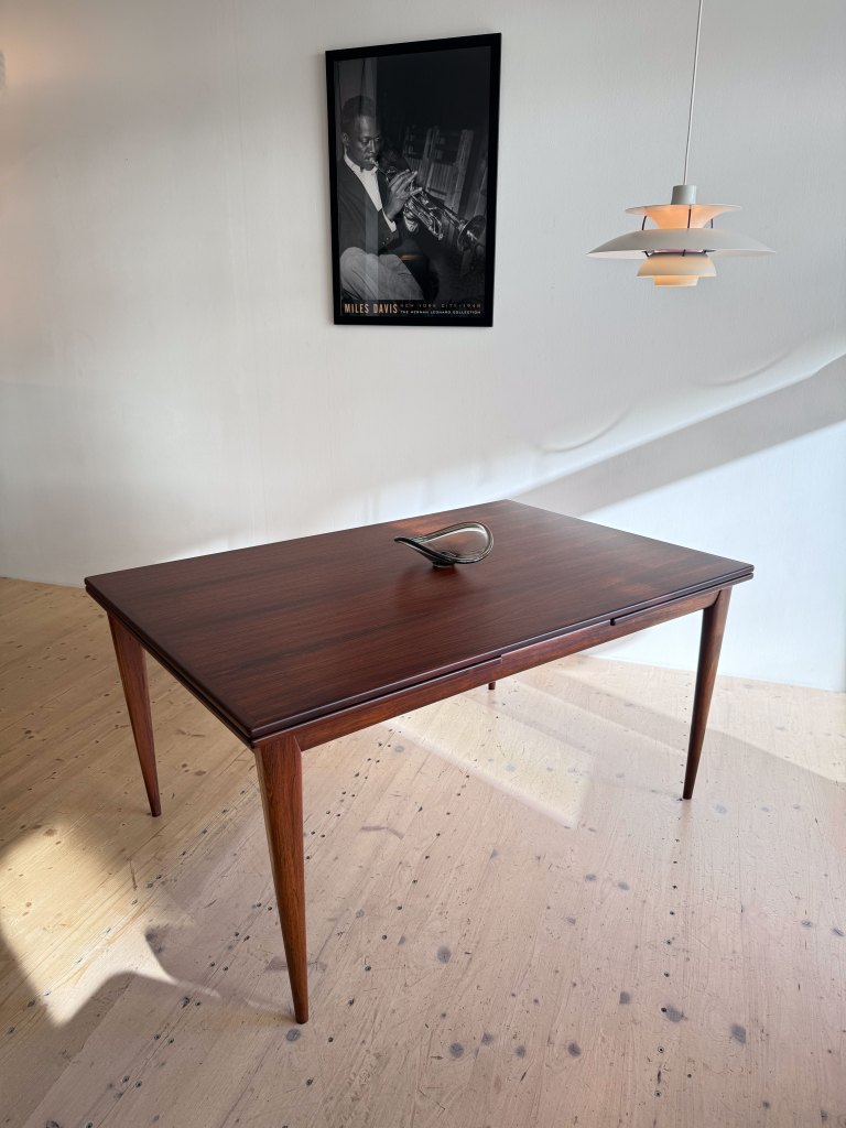 A wooden dining table with tapered legs, a sleek design, and a decorative object on top, set in a well-lit room featuring a framed black and white poster of a musician on the wall and a pendant light hanging above.