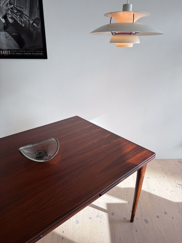 A wooden table with a glossy finish and a minimalist glass bowl on top, illuminated by a stylish pendant lamp, with a framed black and white poster on the wall in the background.