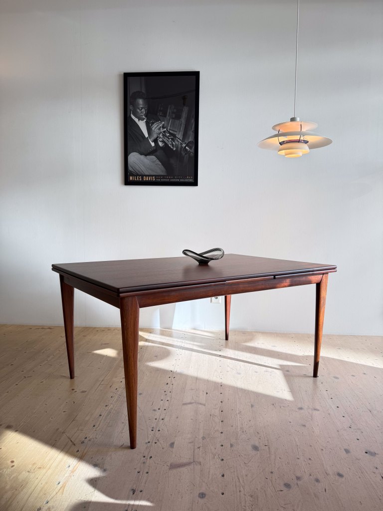 A wooden table with tapered legs and a decorative object on top, positioned in a well-lit room with a poster of a musician on the wall and a pendant light hanging above.