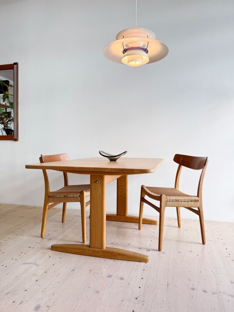 A wooden dining table with two chairs, featuring a decorative bowl on the table and a modern pendant light above, set against a neutral wall and floor.