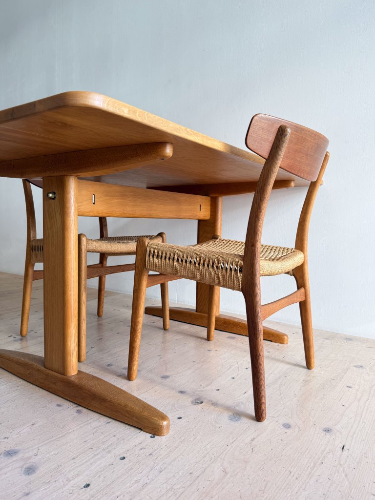 A wooden dining table with rounded edges accompanied by two stylish chairs, one featuring a woven seat, set against a plain wall.