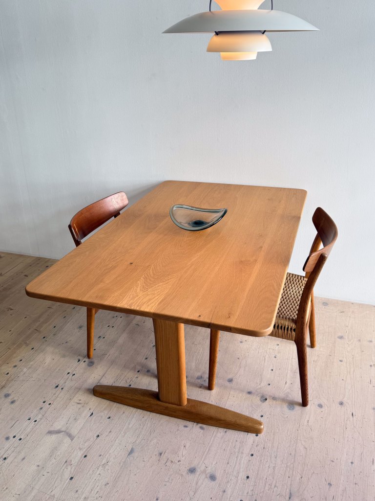 A wooden dining table with two chairs, one with a woven seat, and a decorative bowl on the table, under a pendant light, set against a simple wall.