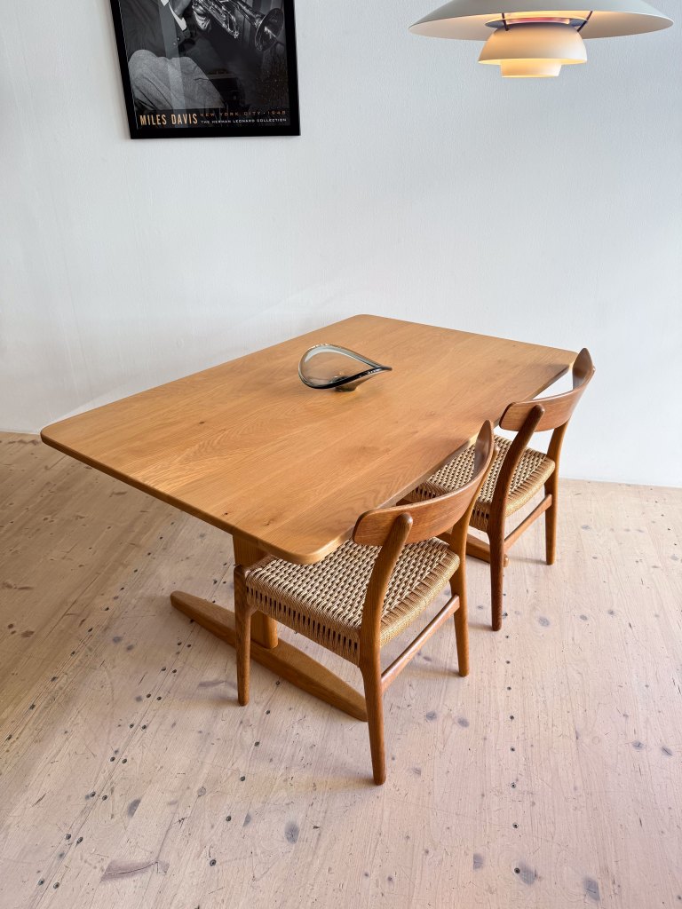 A wooden dining table with two woven chairs, featuring a sleek decorative object in the center and a pendant light hanging above. A black and white poster of Miles Davis is visible in the background.
