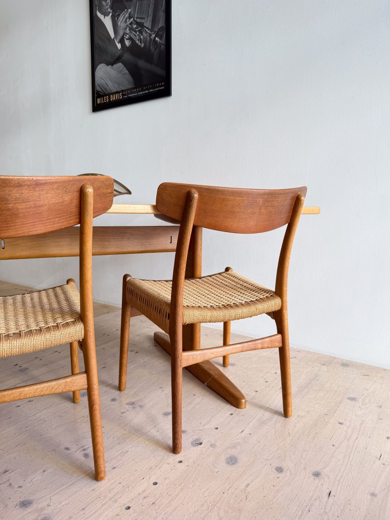 A close-up of two wooden chairs with woven seats placed near a table in a minimalist interior, featuring a poster of a jazz musician on the wall.