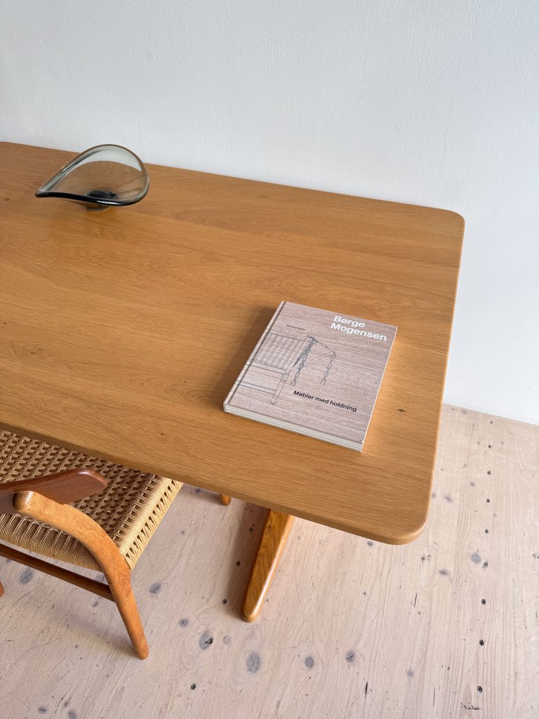 A wooden table with a book titled 'Borge Mogensen' and a glass sculpture on it, featuring a woven chair in the foreground and a light-colored wooden floor.