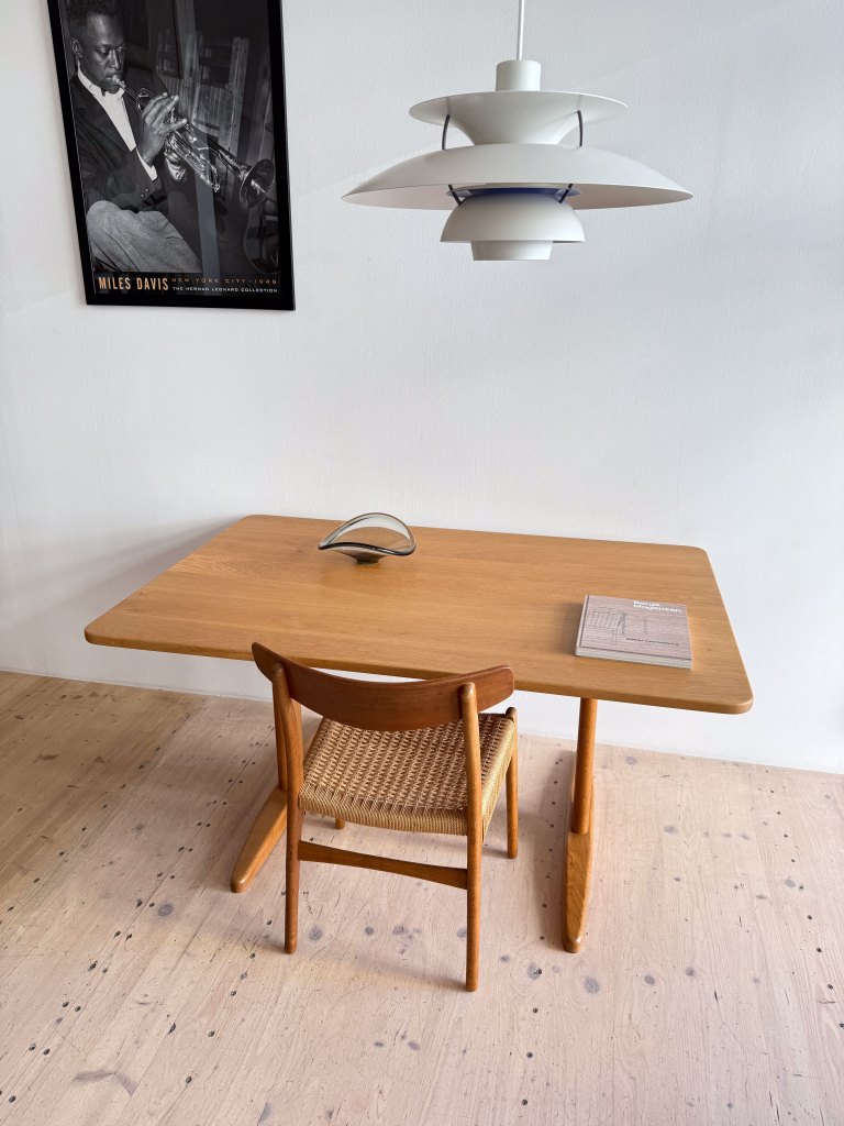 A wooden dining table with a woven chair, a decorative book on the table, and a modern pendant lamp overhead. A framed black and white poster of a jazz musician hangs on the wall.